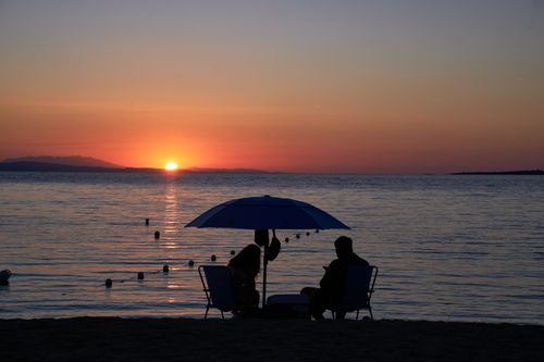 Free Silhouette of a couple enjoying a serene sunset on a Greek beach, under an umbrella. Stock Photo