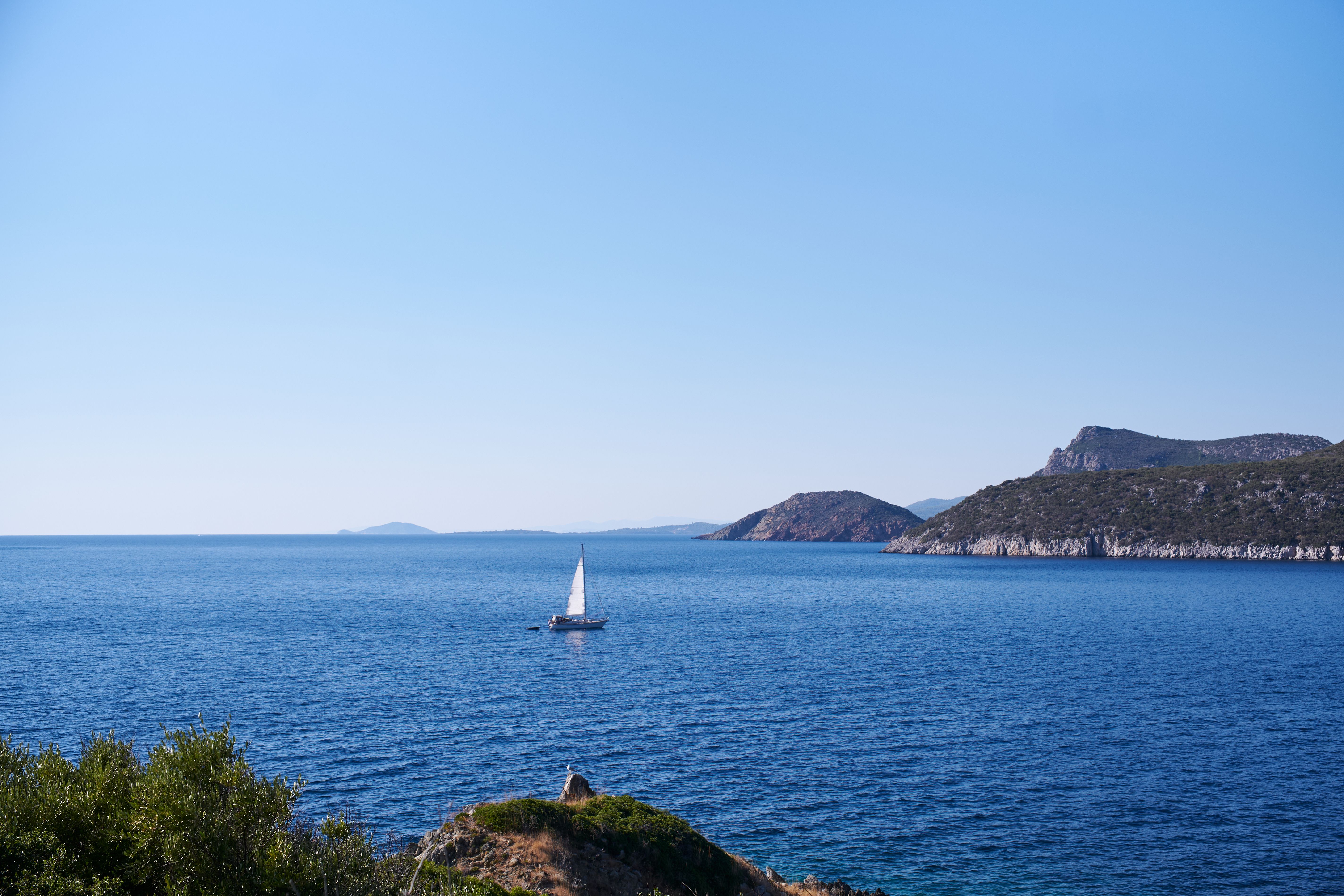 Ultimate All Inclusive Cruise Sailing Cruise to Delos & Rhenia Beaches Review: A Magical Sunset Sailing Adventure in Greece Review 2026 - A serene sailboat glides by the rocky Aegean coastline under clear blue skies.