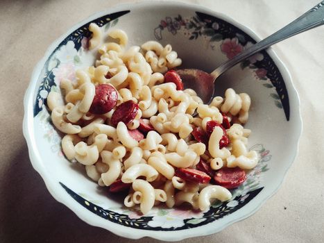 Close-up of creamy macaroni with sliced sausage in a floral design dish, perfect for homestyle cooking visuals.