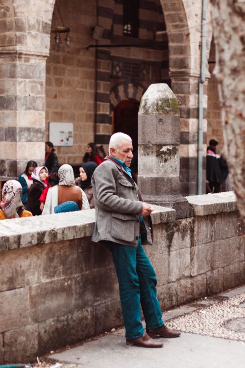 Free An elderly man leans against a historic stone wall in a vibrant courtyard filled with people. Stock Photo