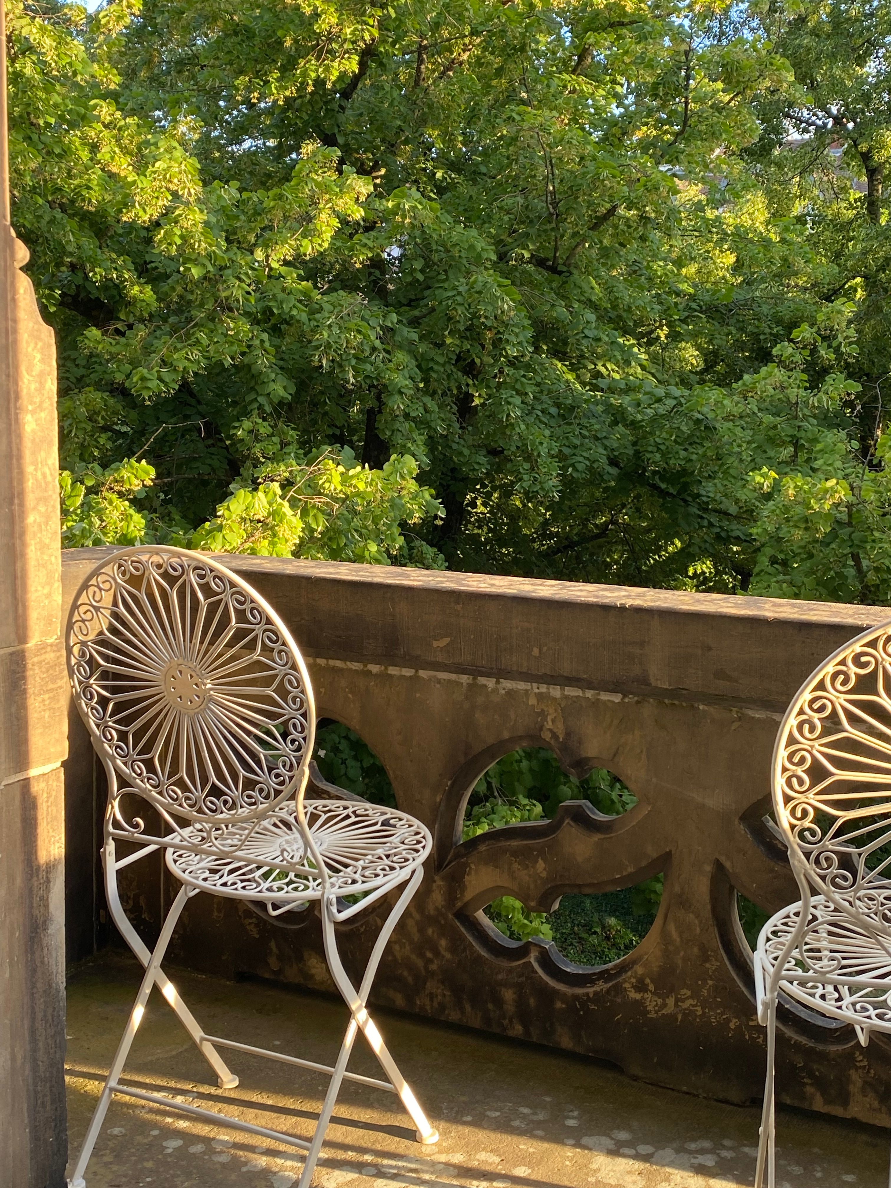 Charming sunlit balcony view with vintage iron chairs overlooking lush green trees.