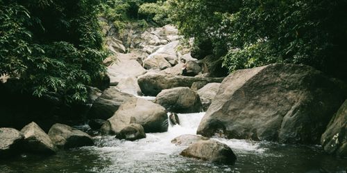 Free A serene waterfall flows over large rocks surrounded by lush greenery in a peaceful forest setting. Stock Photo