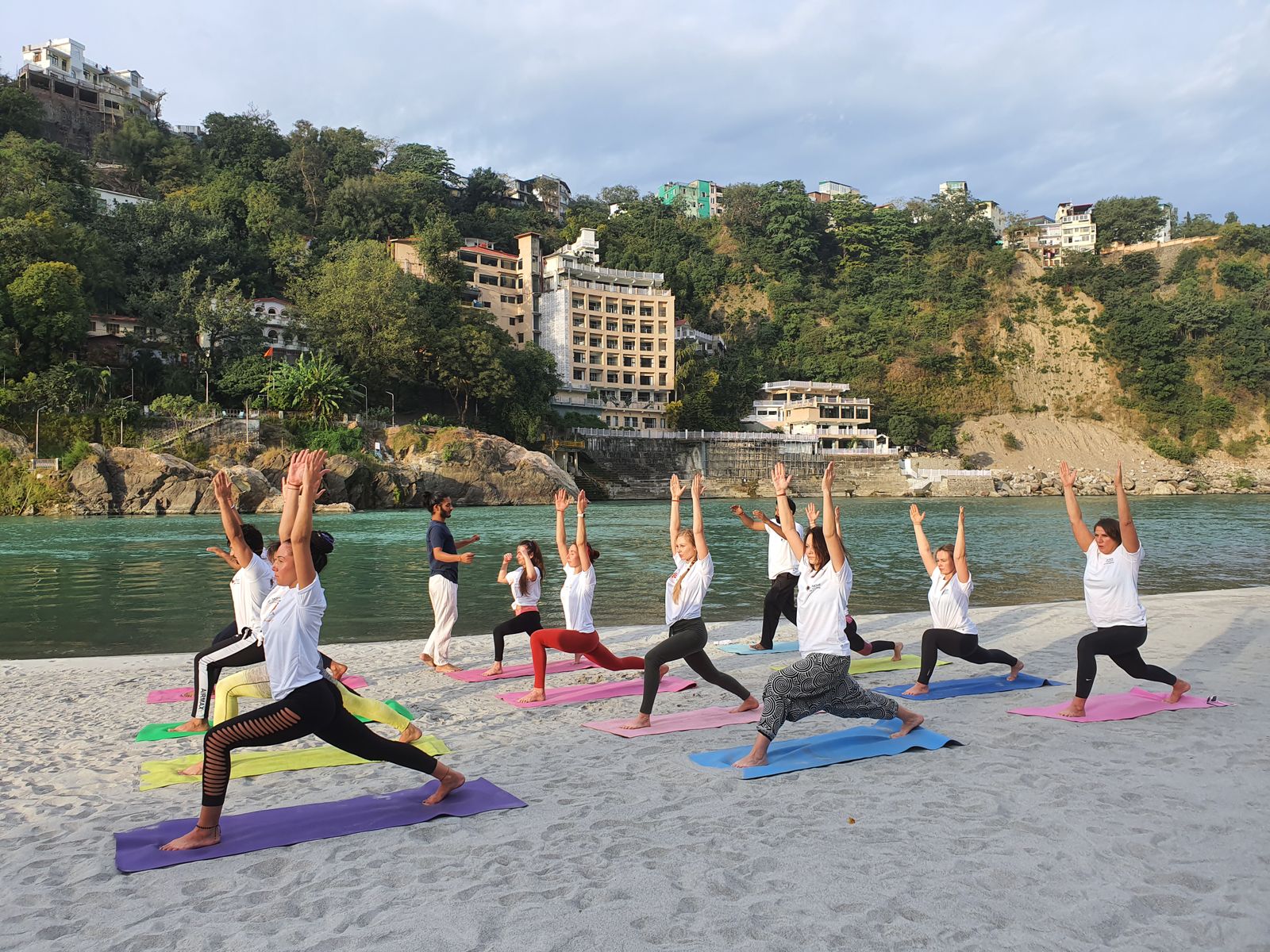 Group of yoga students practicing on mats by the Ganges River in Rishikesh with hills in the background