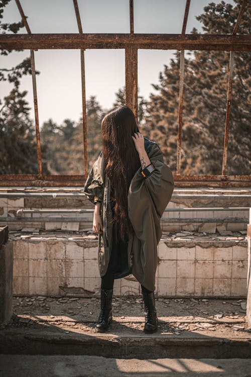 Free Full body of anonymous young woman with long dark hair covering face in stylish warm outfit standing in abandoned shabby building on sunny day Stock Photo