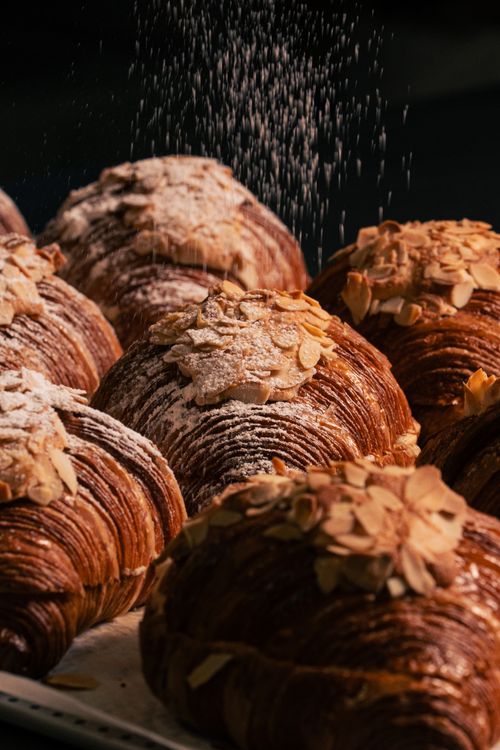 Free Freshly baked artisan almond croissants being dusted with powdered sugar, showcasing delicate layers and texture. Stock Photo