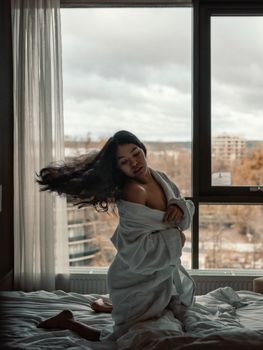 Elegant woman in a bathrobe posing by a window in a comfortable bedroom setting.