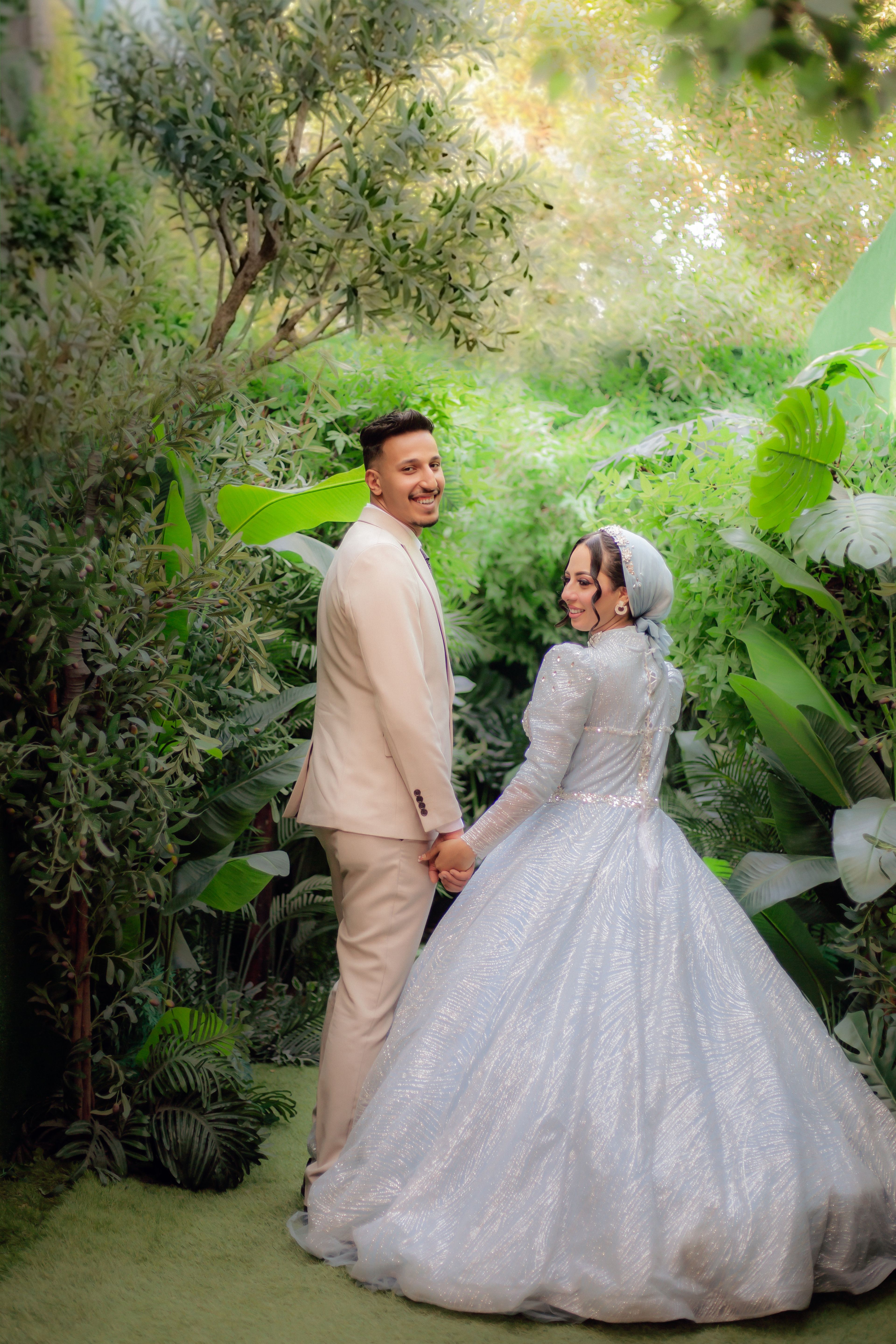 A joyful couple in wedding attire posing amidst vibrant greenery in an outdoor setting.