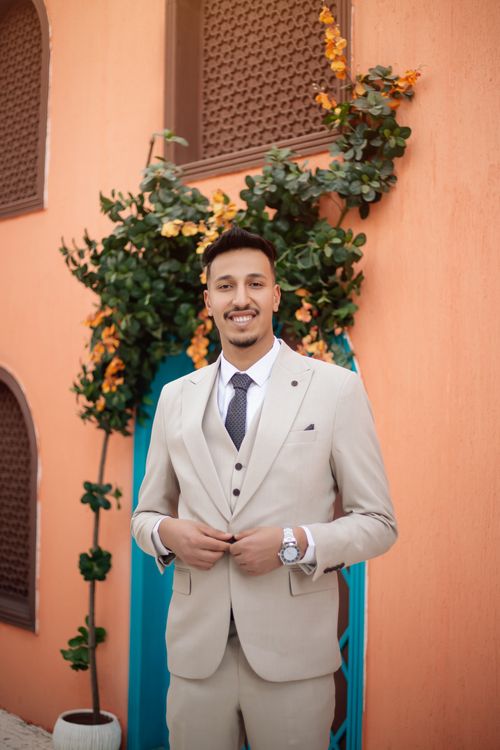 Free A groom in a beige suit standing outdoors against a decorative wall with greenery. Stock Photo