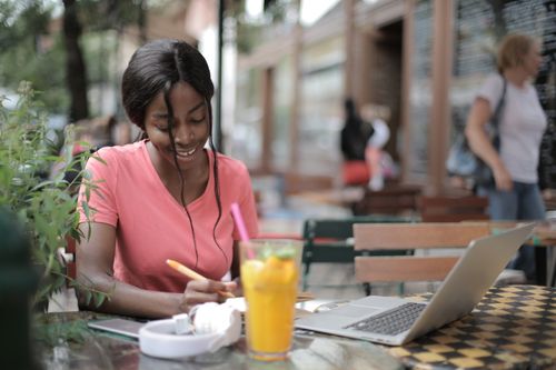 Free Smiling woman in pink shirt writing at a café with laptop and juice. Stock Photo