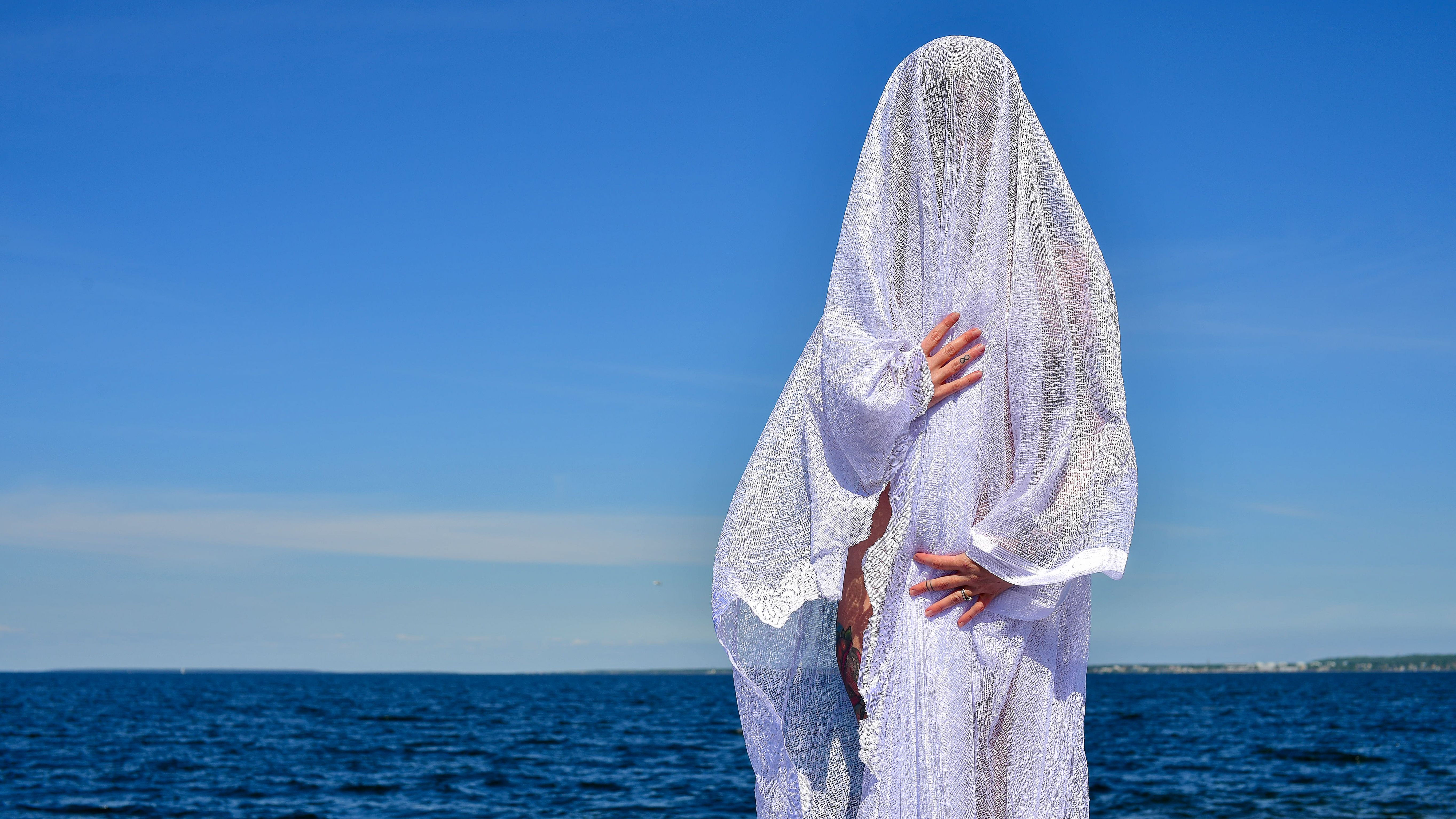 A mysterious figure draped in white fabric stands by the sea under a clear blue sky in Tallinn, Estonia.