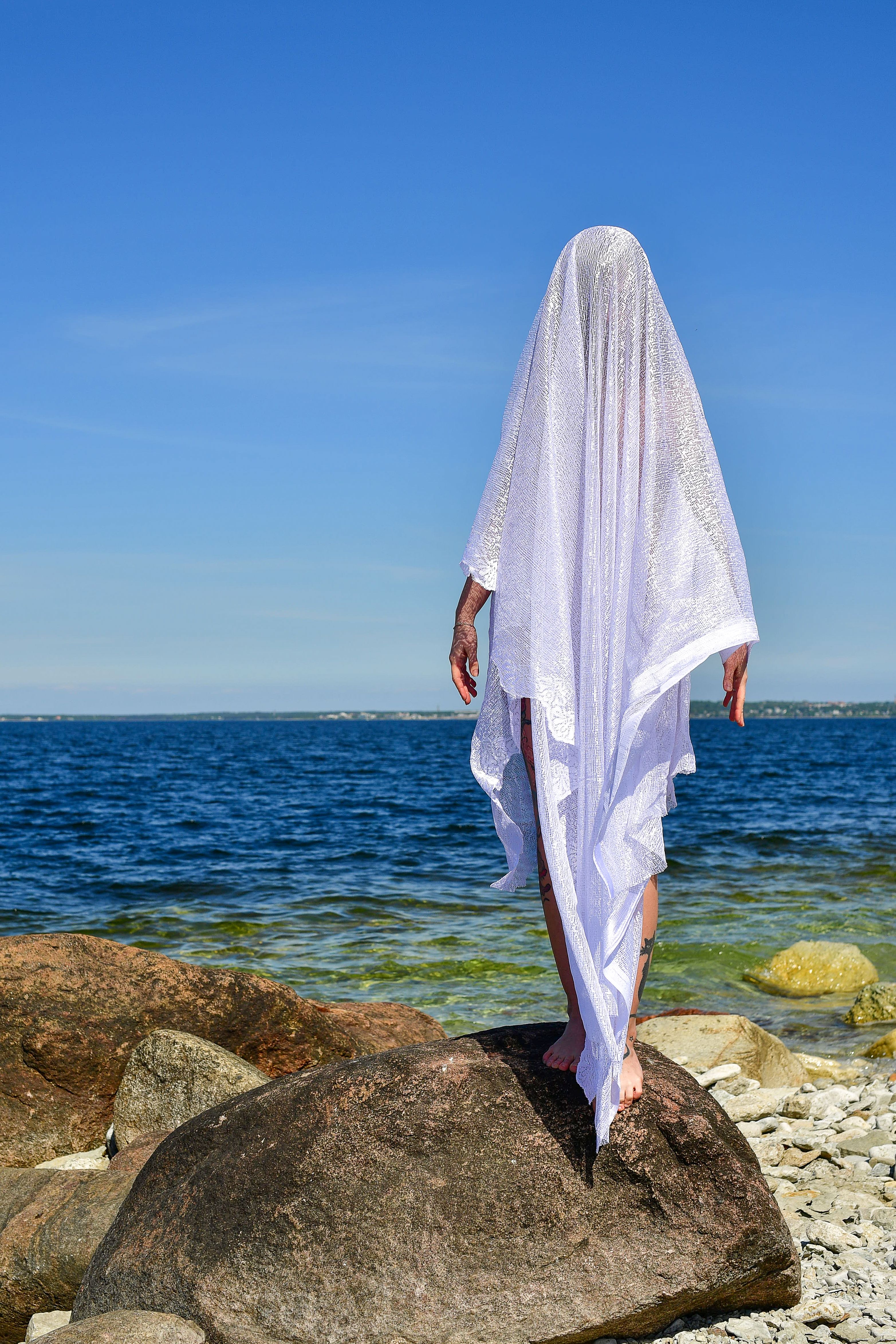 A mysterious figure draped in white fabric stands on a rocky Estonian beach by the sea.
