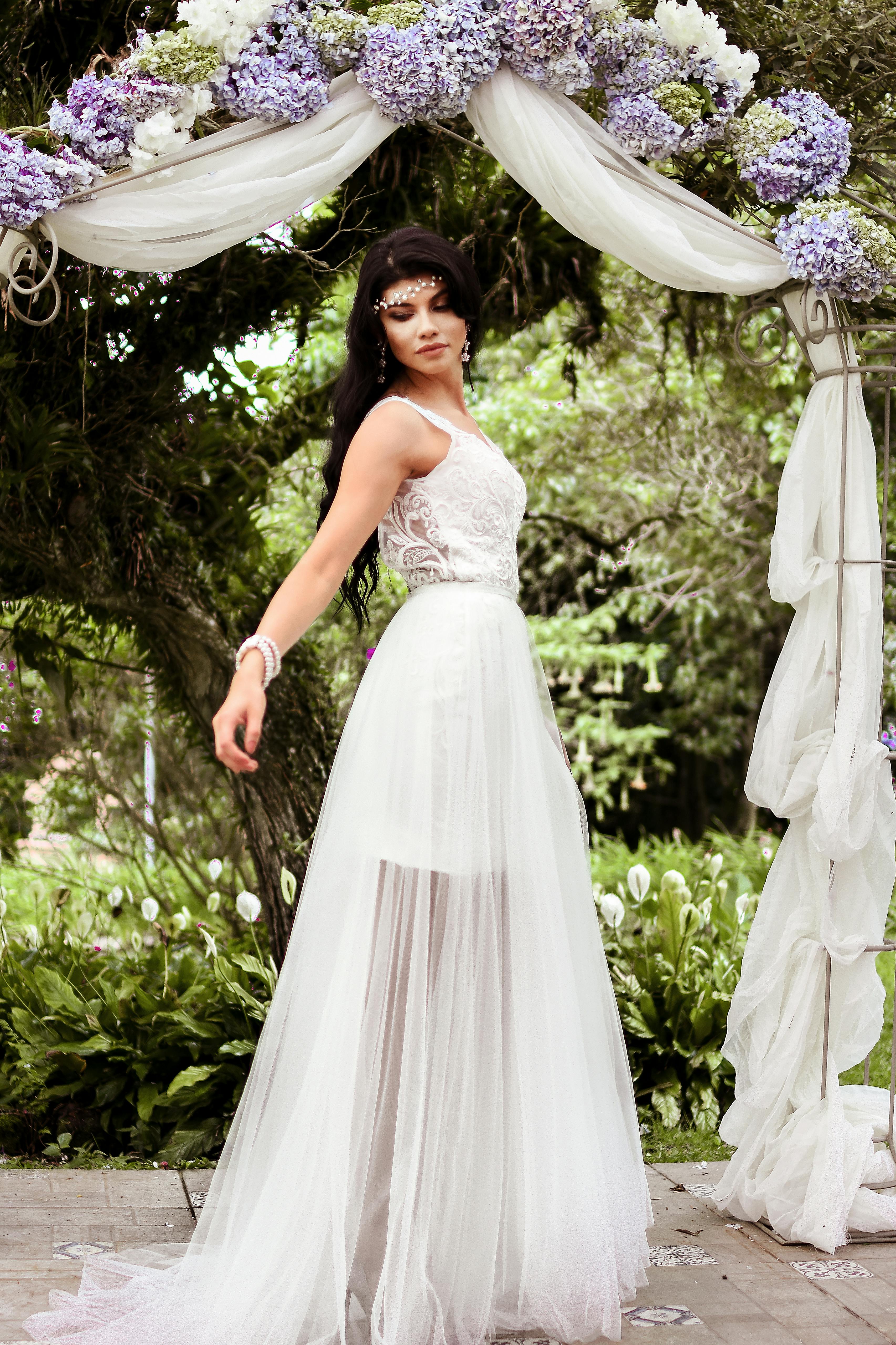A beautiful bride poses in a stunning wedding gown under a floral arch outdoors.