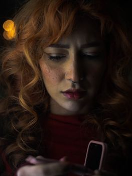 Moody portrait of a woman with curly red hair and a nose ring, focusing intently indoors.
