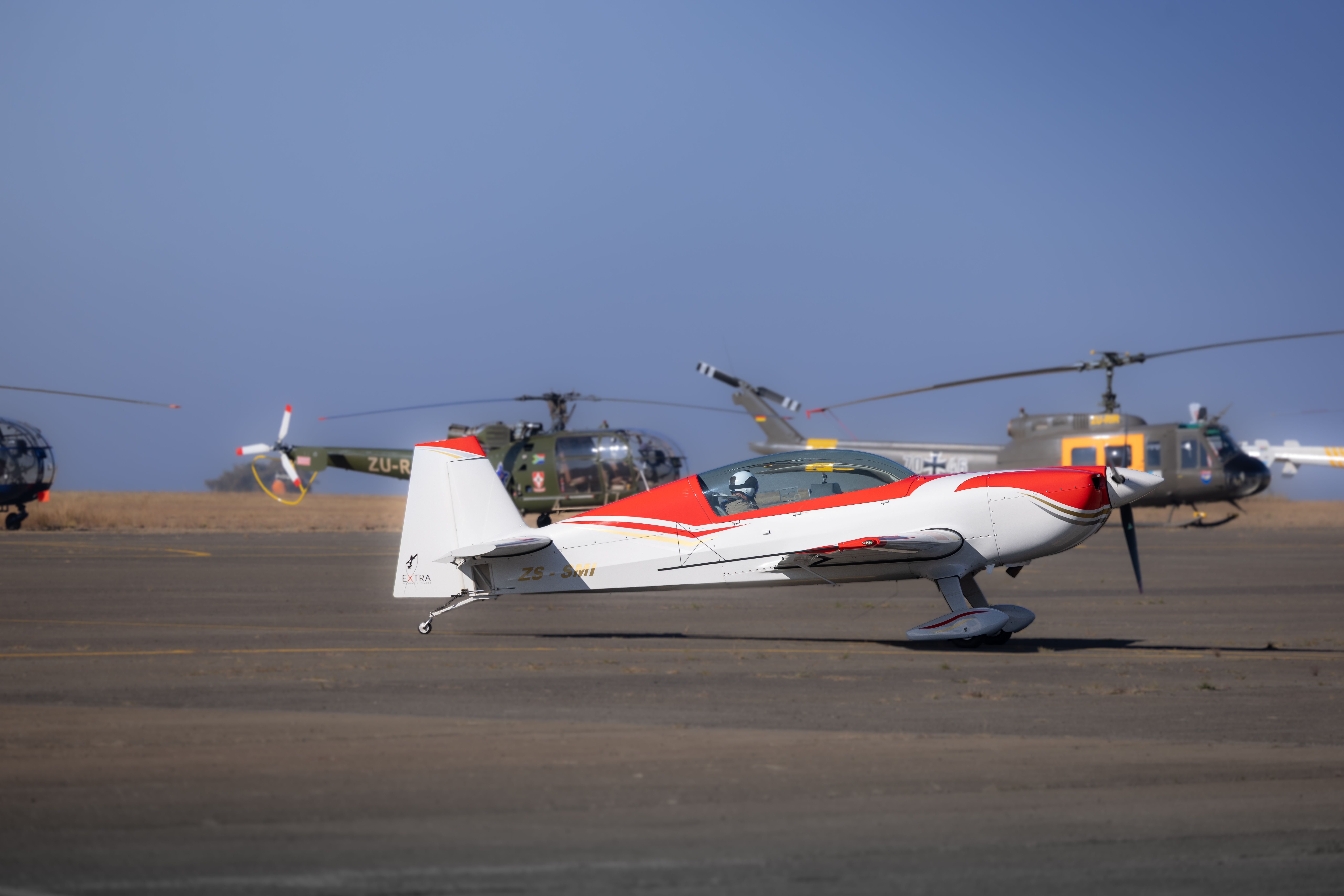 A white and red aerobatic plane parked on an airfield with helicopters in the background.