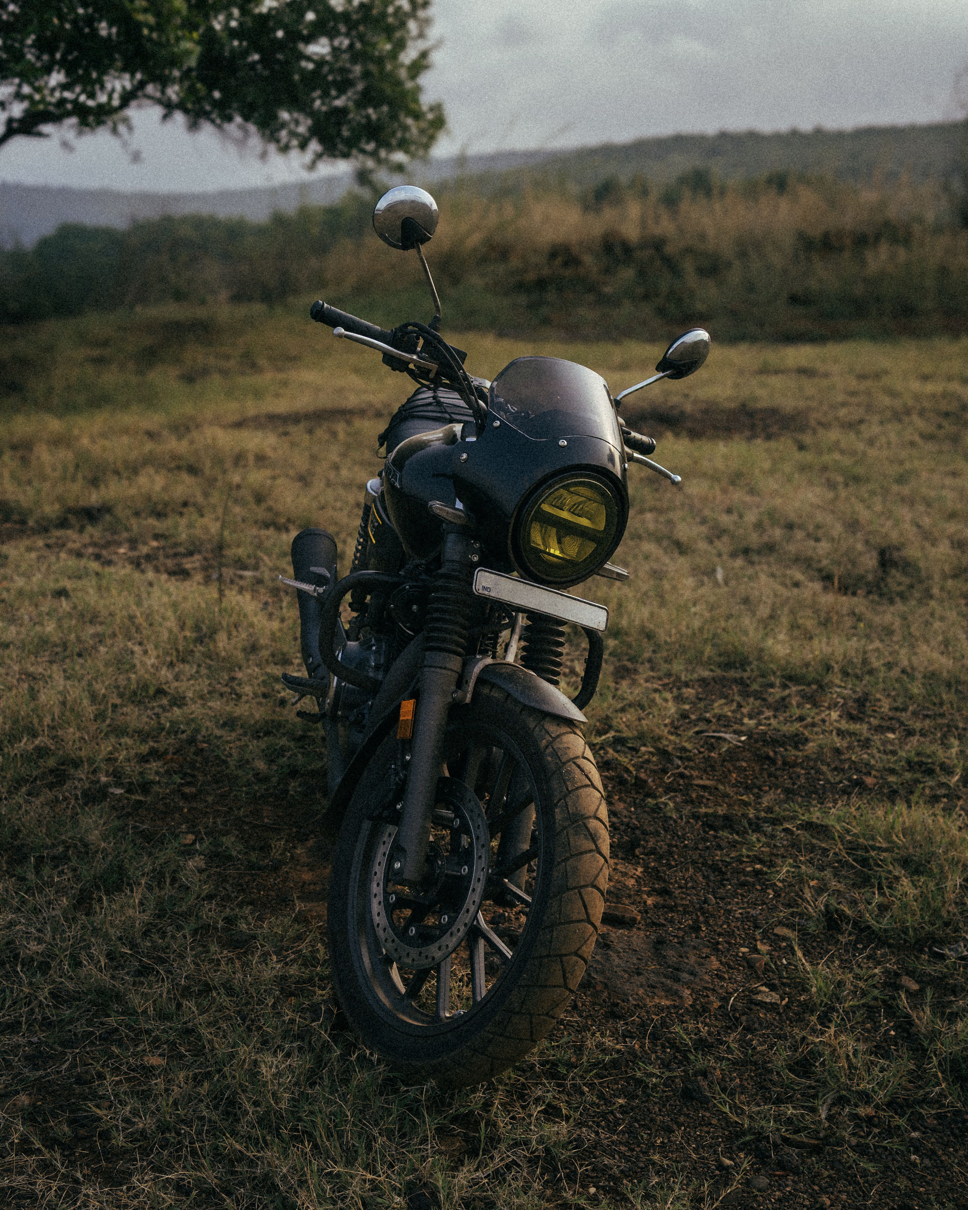 A classic vintage motorcycle parked on grassy terrain in rural India, under a cloudy sky.