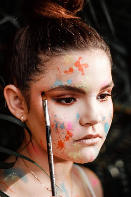 Free Close-up portrait of a woman with colorful face paint and a paintbrush, capturing a thoughtful expression. Stock Photo