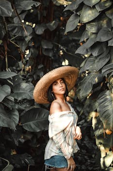 A stylish woman in denim shorts and straw hat poses elegantly amidst lush green foliage.