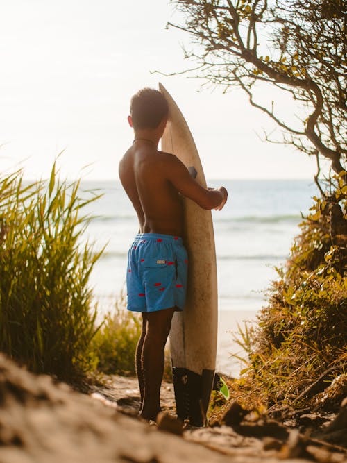 Topless Man in Blue Shorts While Holding White Surfboard