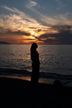 A serene silhouette of a woman standing on a beach during a vibrant sunset over the ocean.