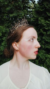 Side profile of a woman wearing a crown, set against lush greenery. Elegant and regal.