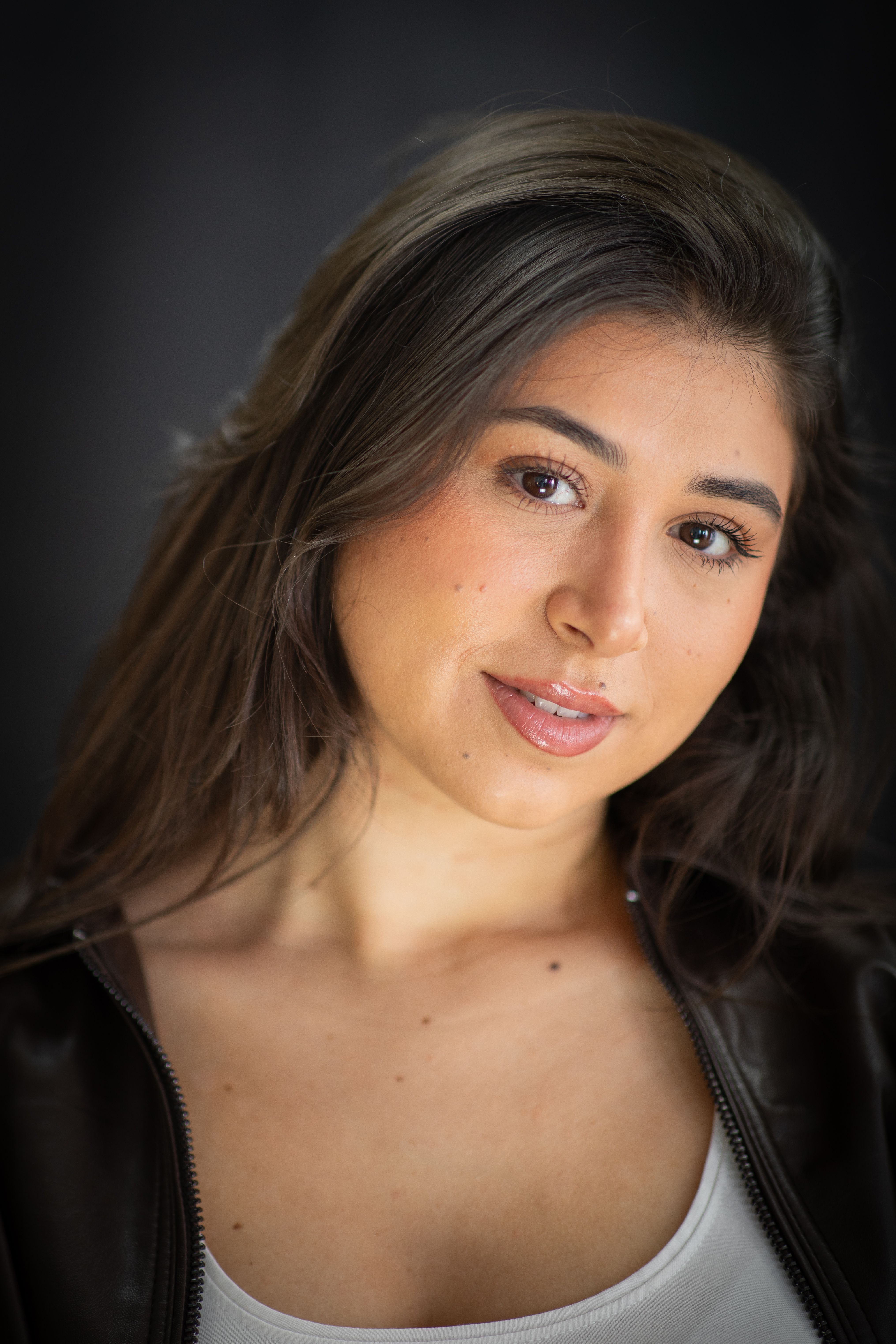 Close-up portrait of a young woman with a friendly smile wearing a leather jacket against a dark background.