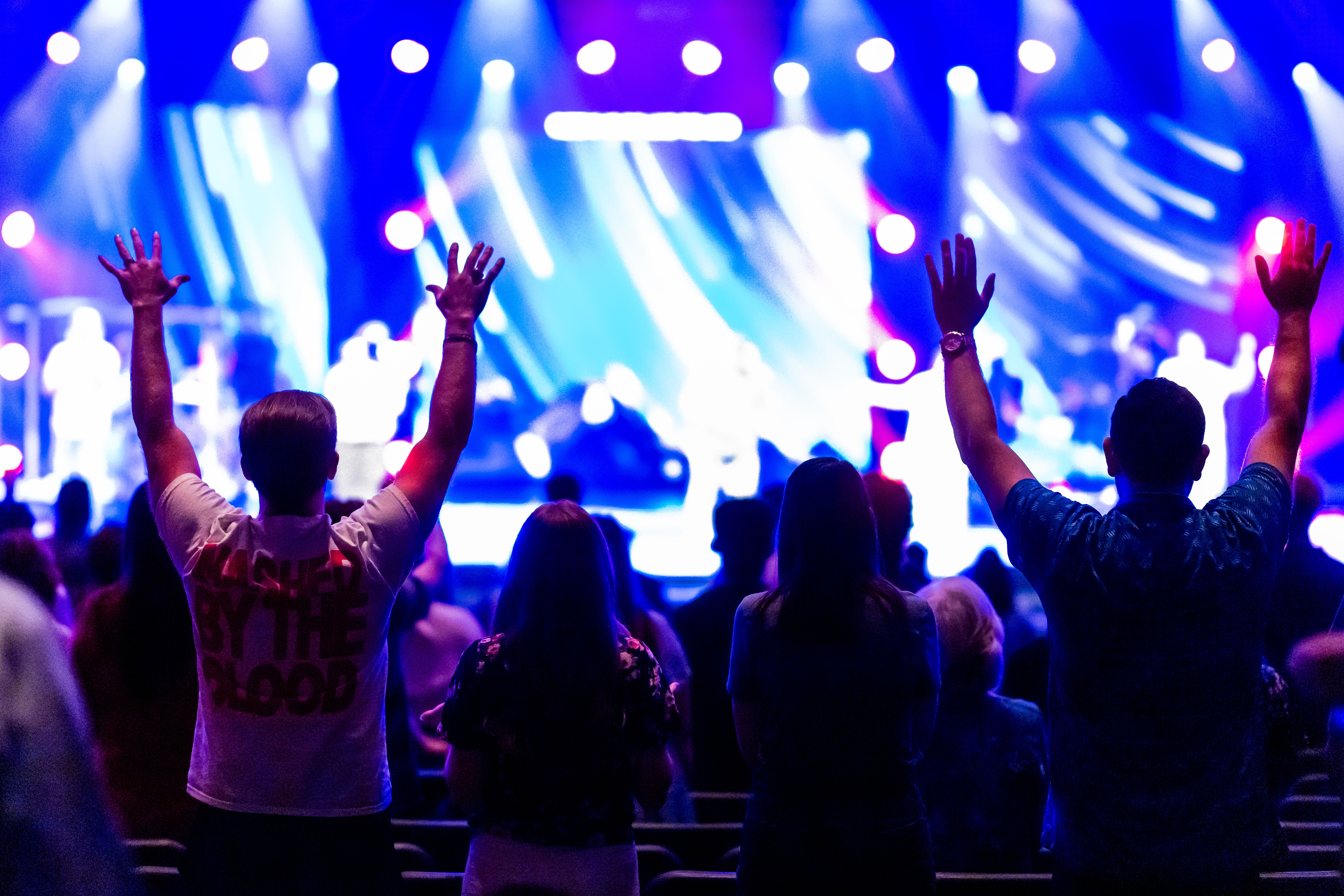 Vibrant crowd at an indoor concert with colorful stage lights and raised hands.