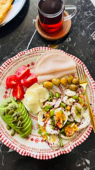 Delicious and nutritious breakfast plate featuring avocado, eggs, turkey slices, olives, and tomato with a side of tea.