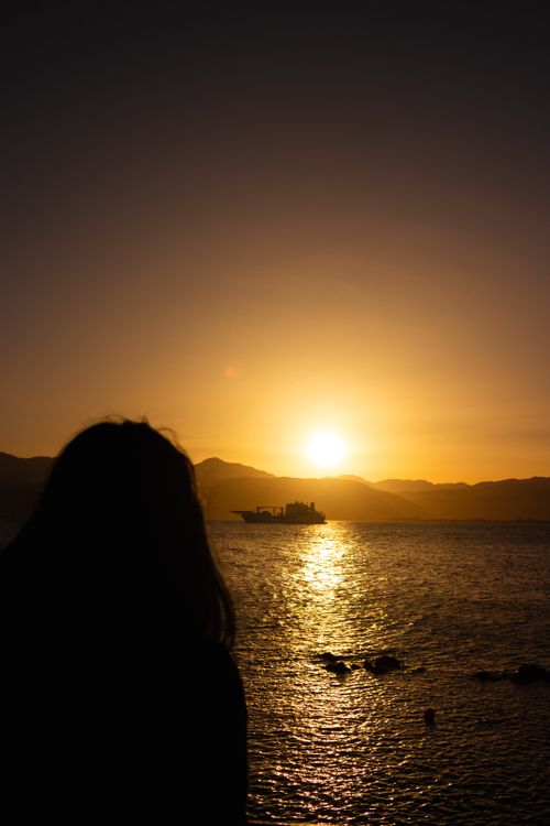 Free A serene silhouette of a woman watching the sunset over the sea in Greece, with a distant ship on the horizon. Stock Photo