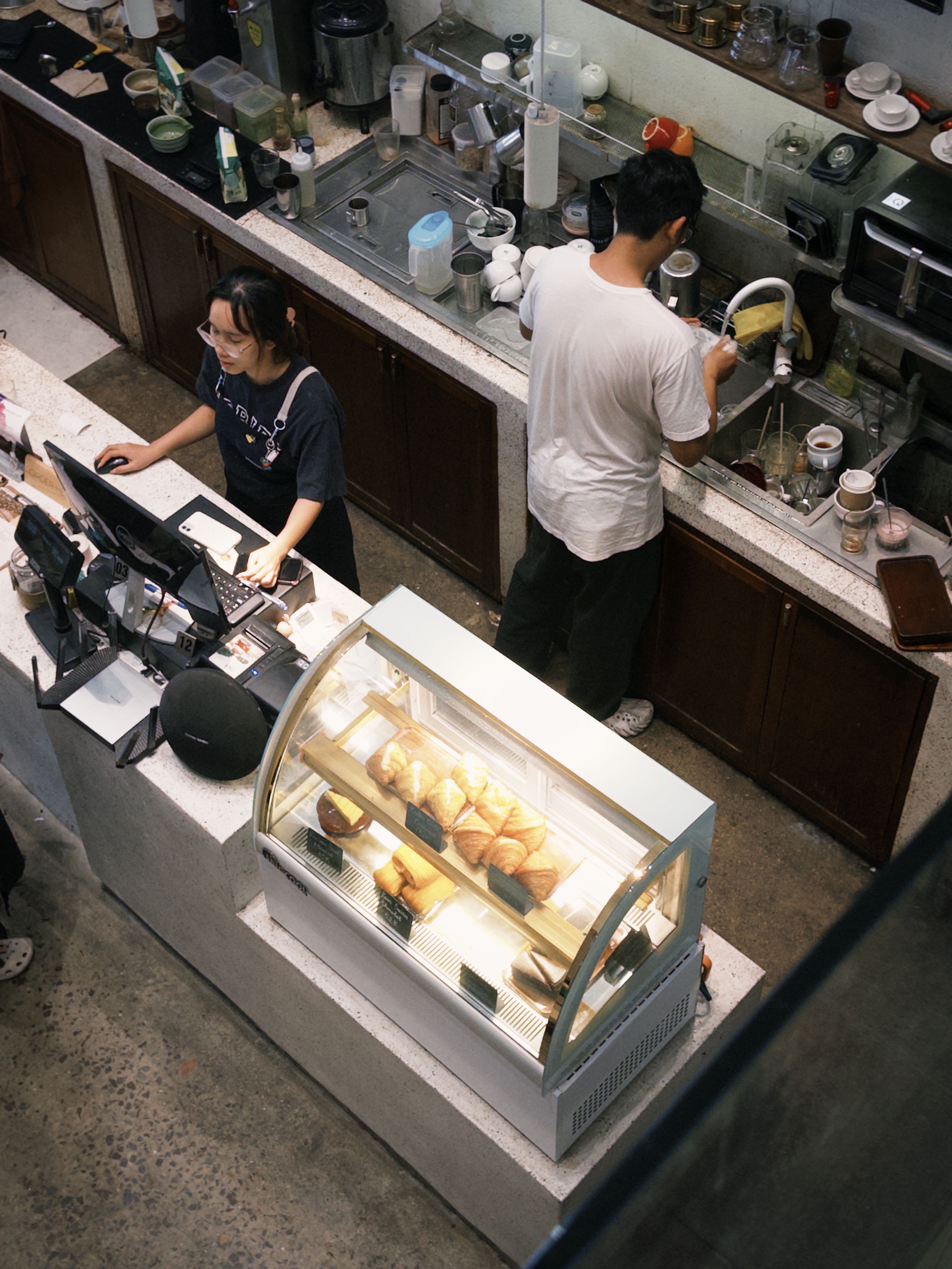 Top view of a bustling coffee shop counter with two baristas preparing beverages and baked goods on display.