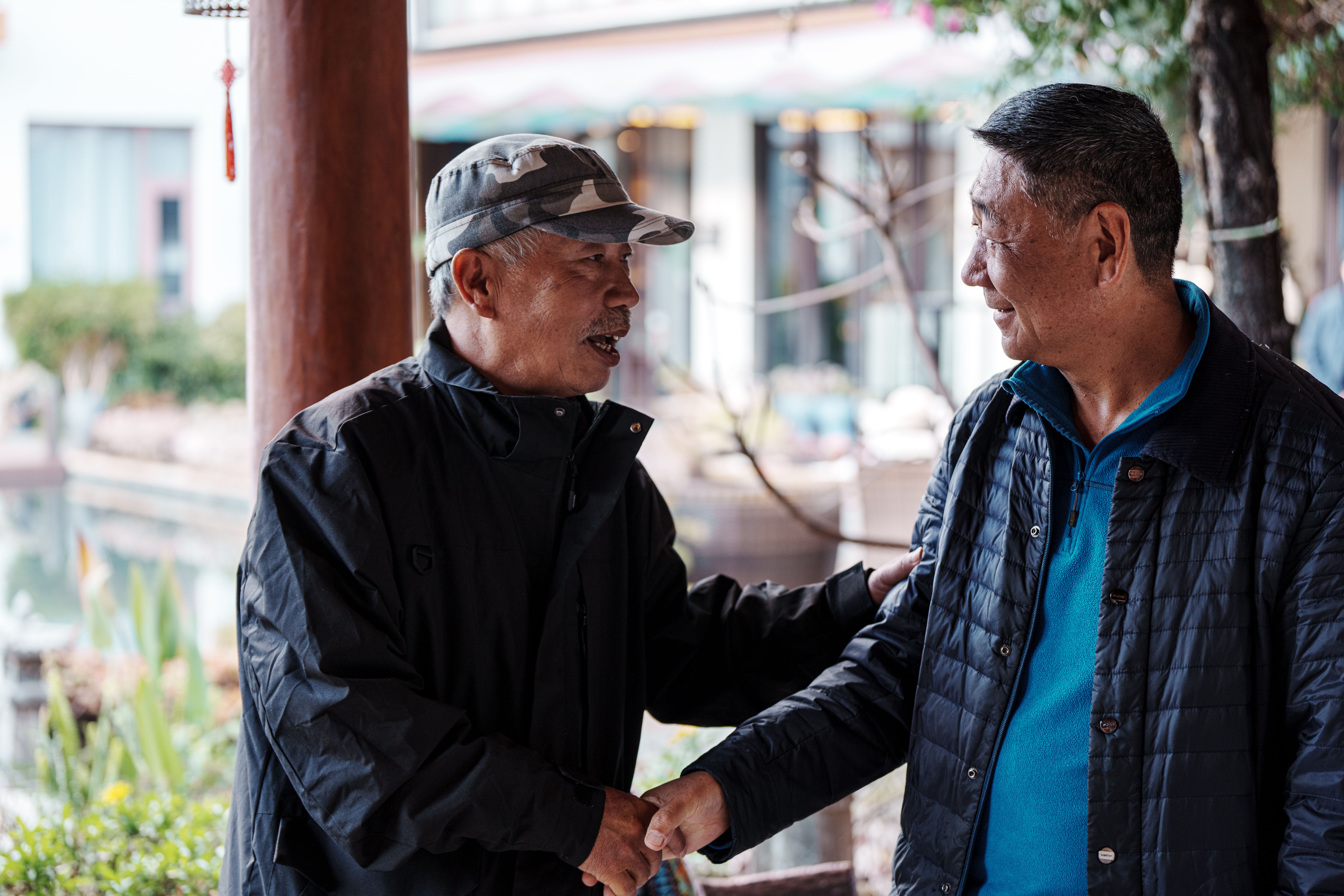 Two older men greet each other warmly outside in a relaxed setting.