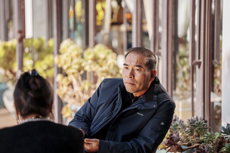 Two adults engaged in a relaxed conversation in a sunny outdoor cafe with plants.