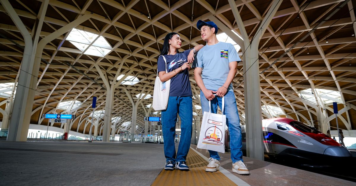 A young couple smiling and posing at a modern high-speed train station with a sleek bullet train in Jakarta, Indonesia.