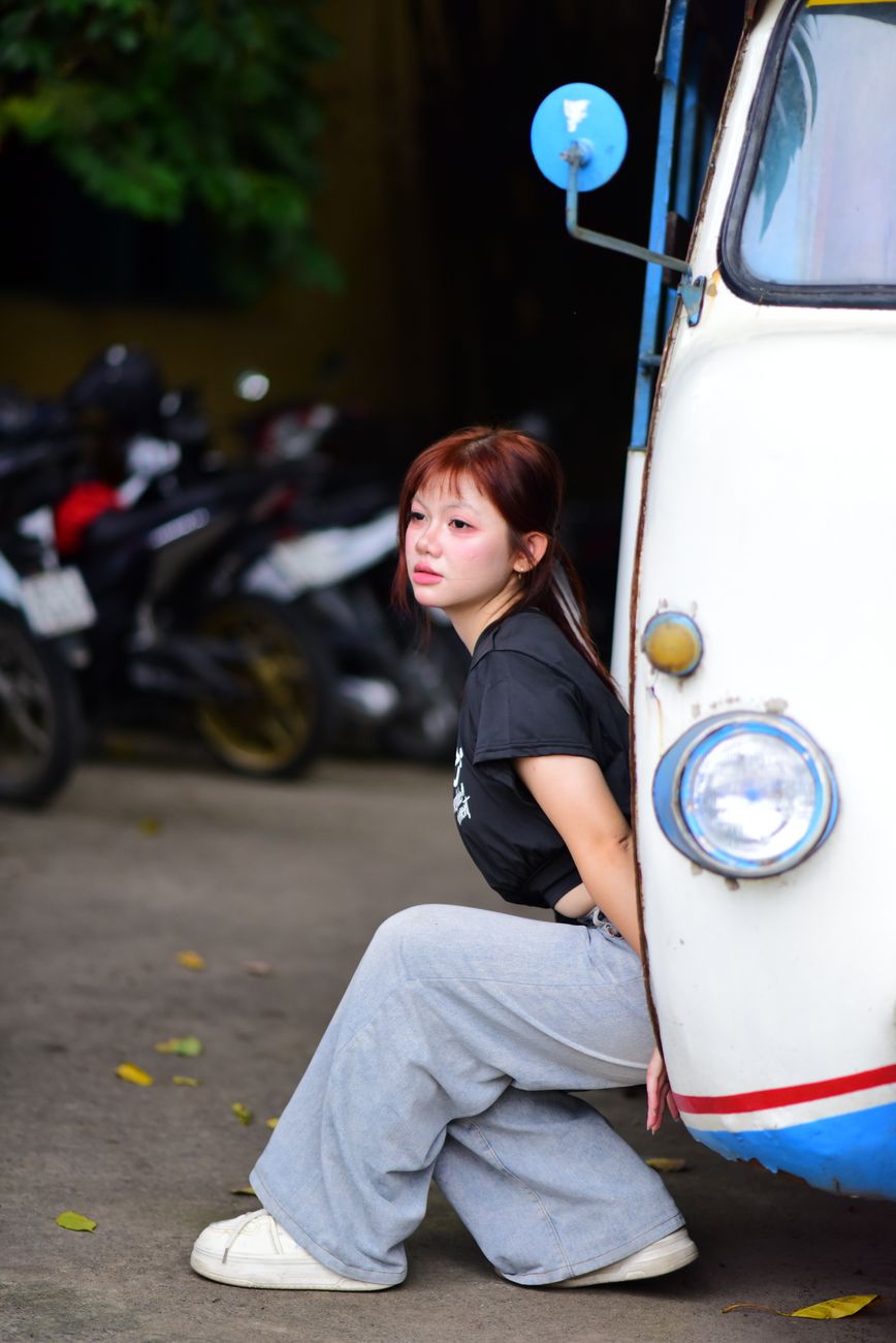 A young woman in casual attire poses thoughtfully by a colorful vintage vehicle outdoors.