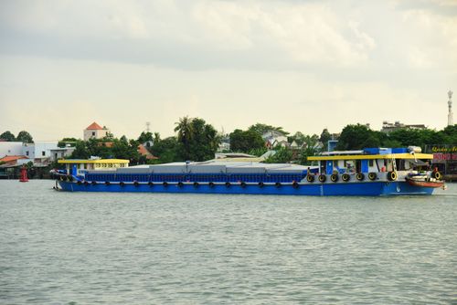 Free A large blue cargo ship travels along a serene river with a town in the background, under a cloudy sky. Stock Photo