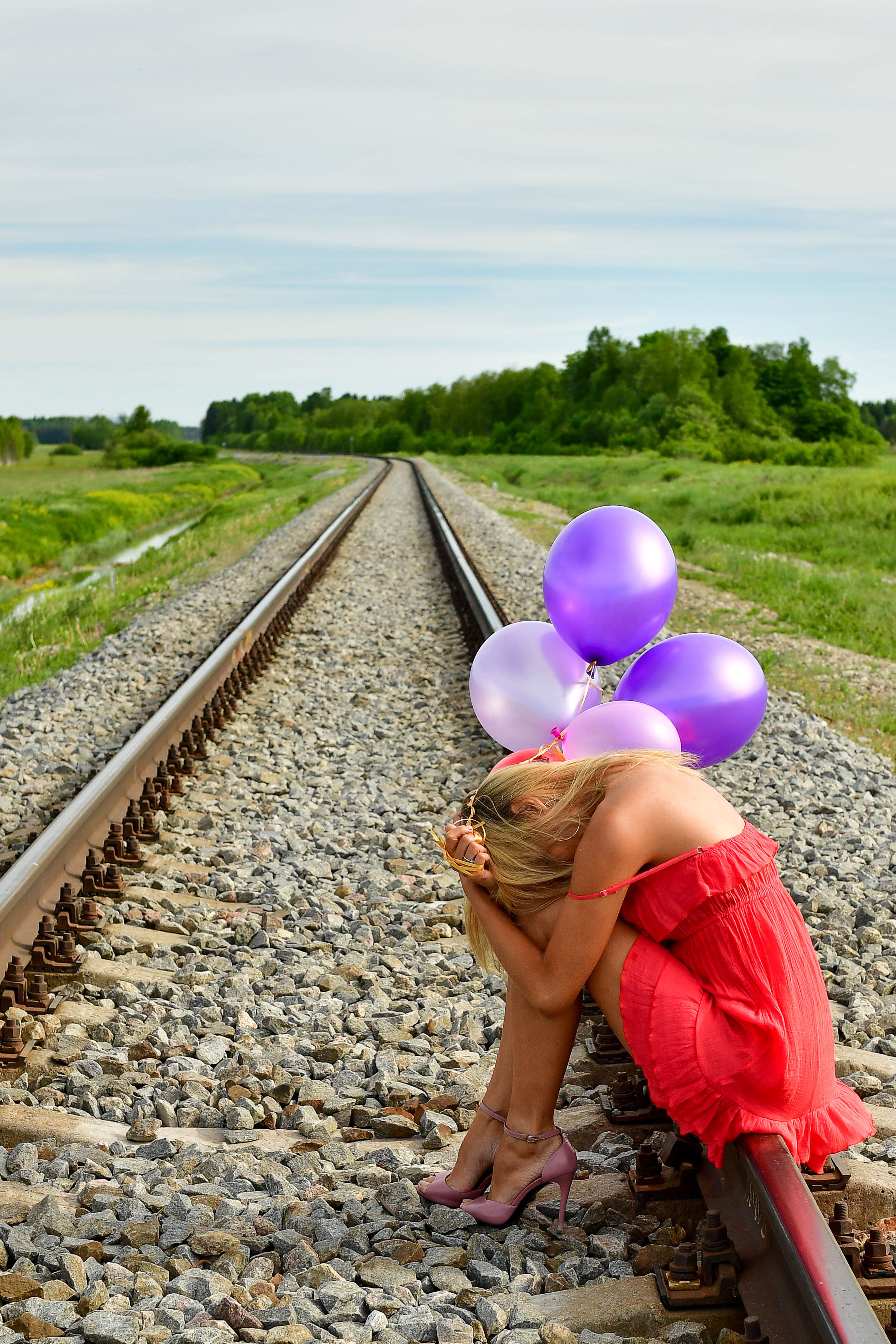Woman in red dress with purple balloons portraying solitude on railway tracks.
