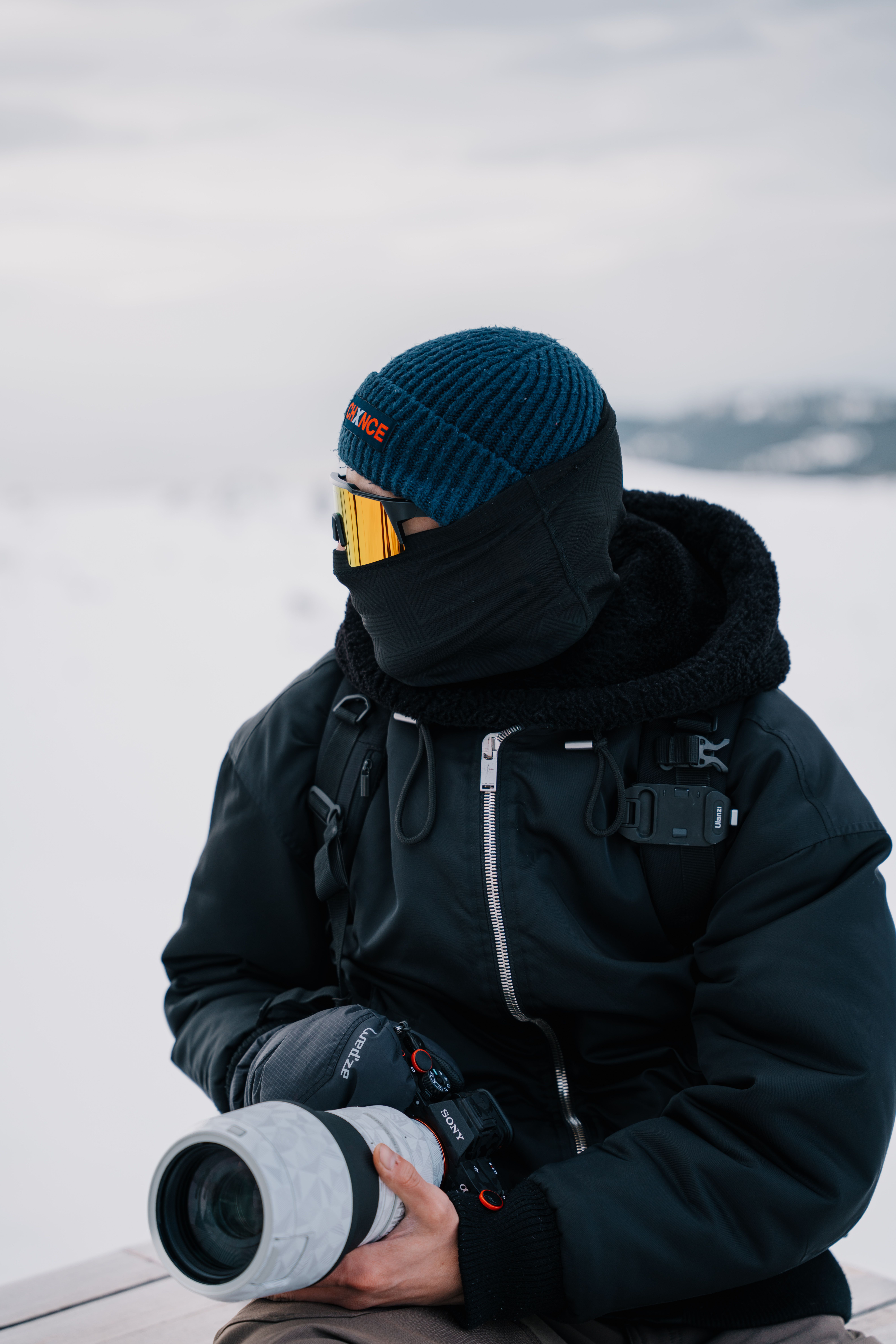 A bundled-up photographer holds a telephoto lens in a snowy outdoor setting.
