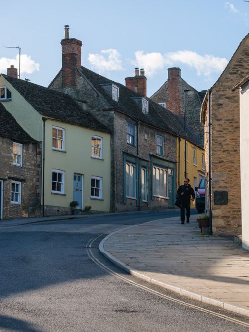 Free Picturesque street scene in Malmesbury, England with historic stone houses and a lone walker. Stock Photo