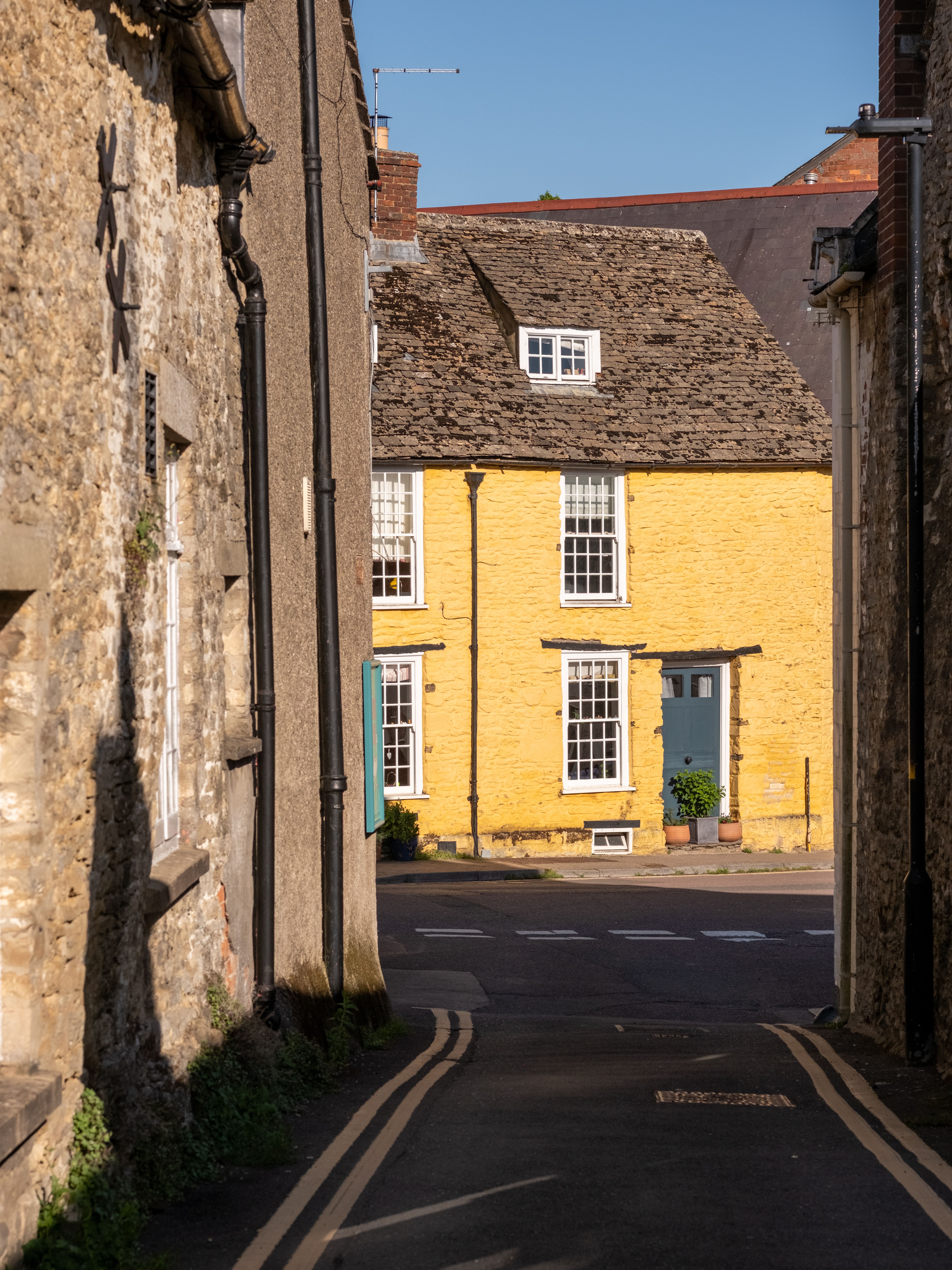 A picturesque yellow cottage on a quaint street in Malmesbury, England, under clear blue skies.
