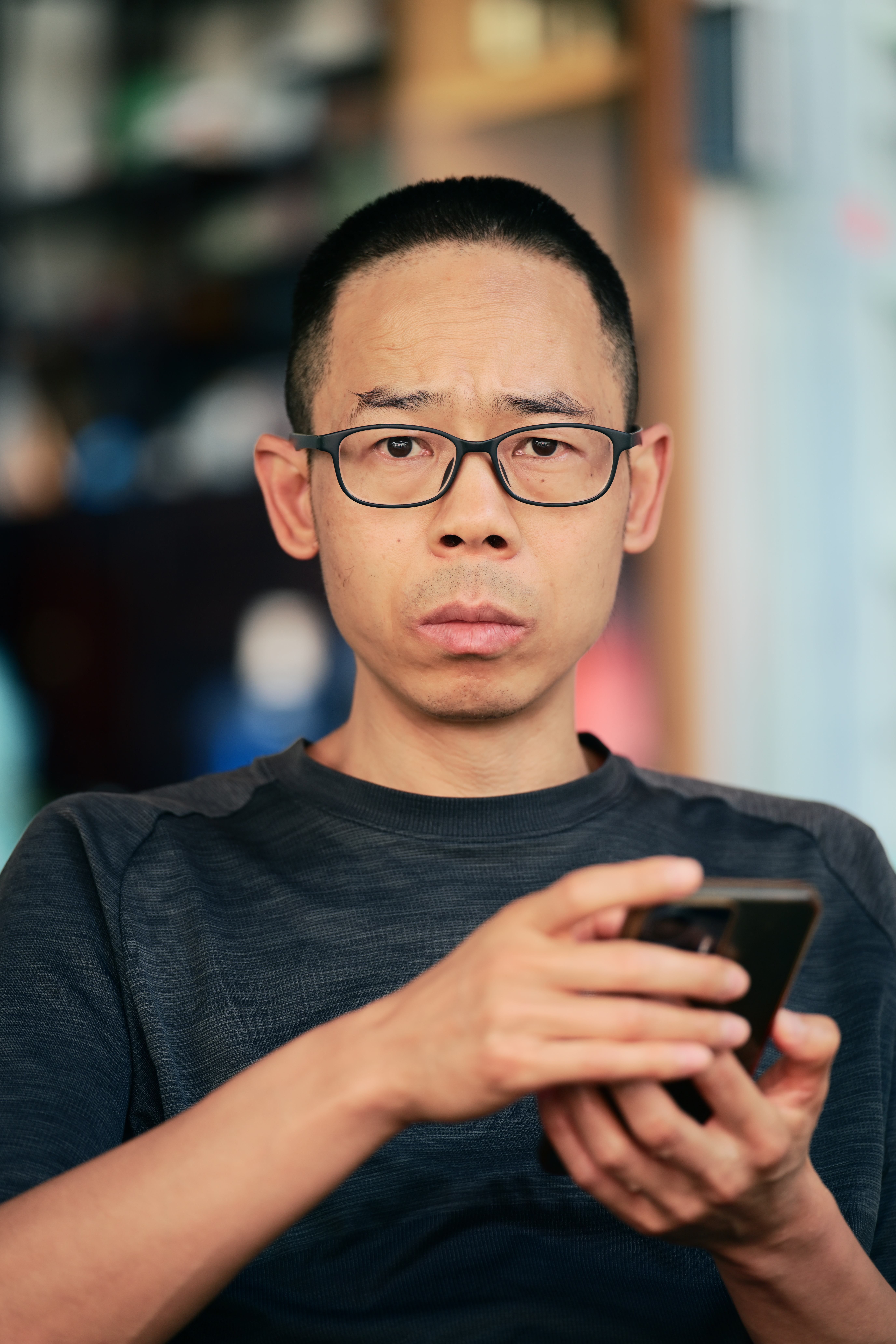 An adult man with glasses holding a smartphone, looking at the camera indoors.