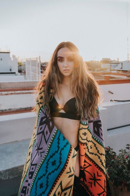 Portrait of a woman in a black bra, wrapped in a colorful blanket on a rooftop at sunset.