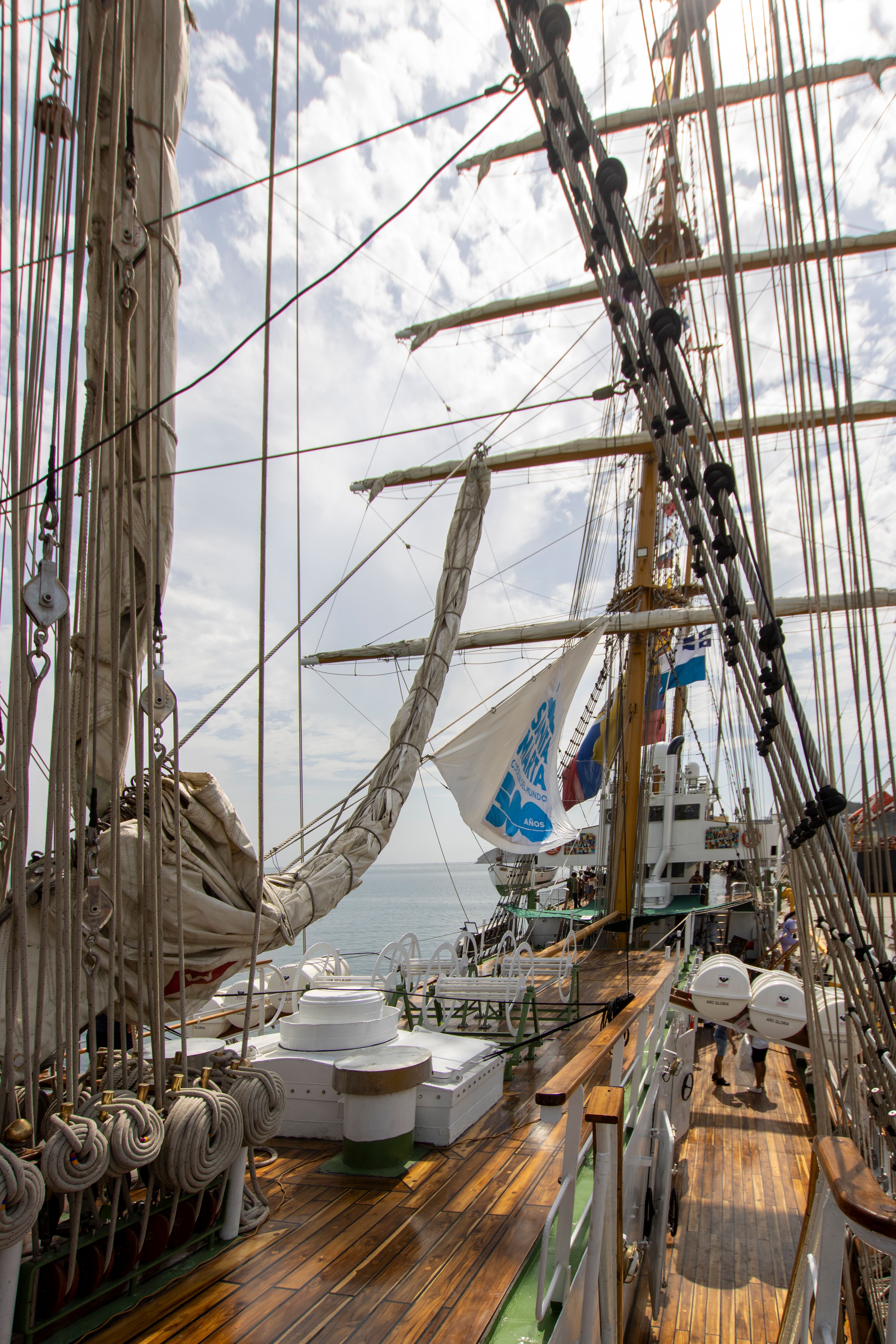 View of a sailing ship's deck featuring rigging and flags against a cloudy sky.