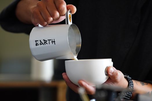 A barista skillfully pours milk into a coffee cup. The milk jug is labeled 'Earth'.
