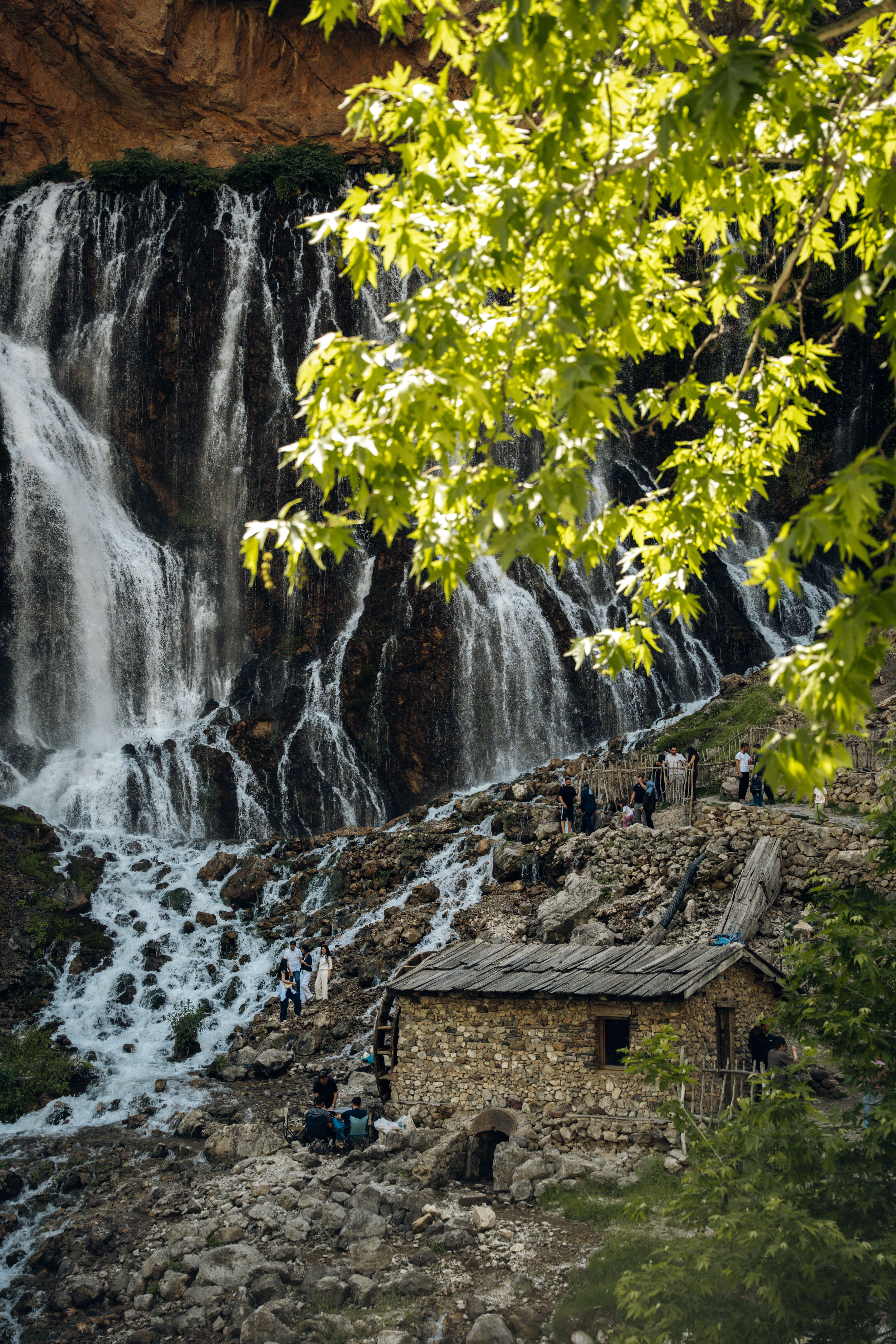 Stone cabin nestled by a stunning waterfall in lush greenery.