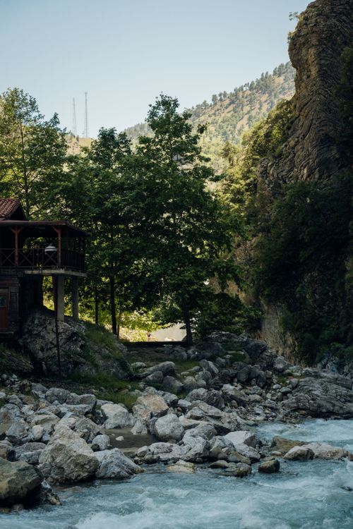 Free Scenic view of a riverside cabin surrounded by lush trees and rocky terrain in a mountainous landscape. Stock Photo