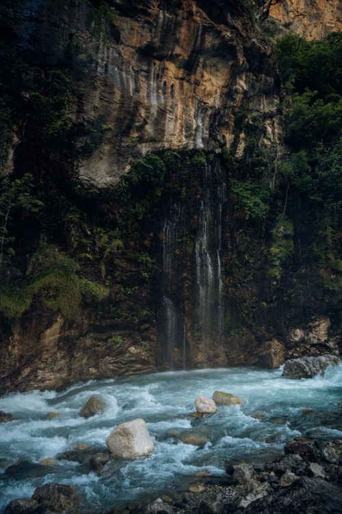 Free A stunning waterfall cascades down a rocky cliff into a vibrant blue stream surrounded by lush greenery. Stock Photo