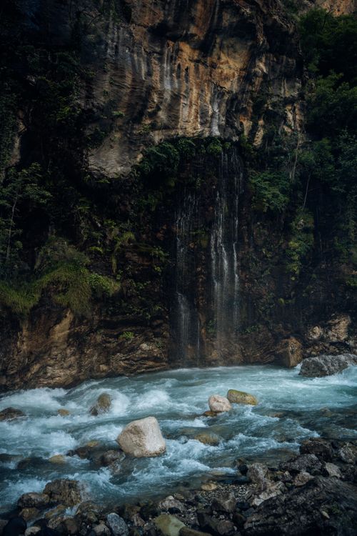 Free A tranquil waterfall flows down a rocky cliff into a clear, bubbling stream in a lush forest setting. Stock Photo