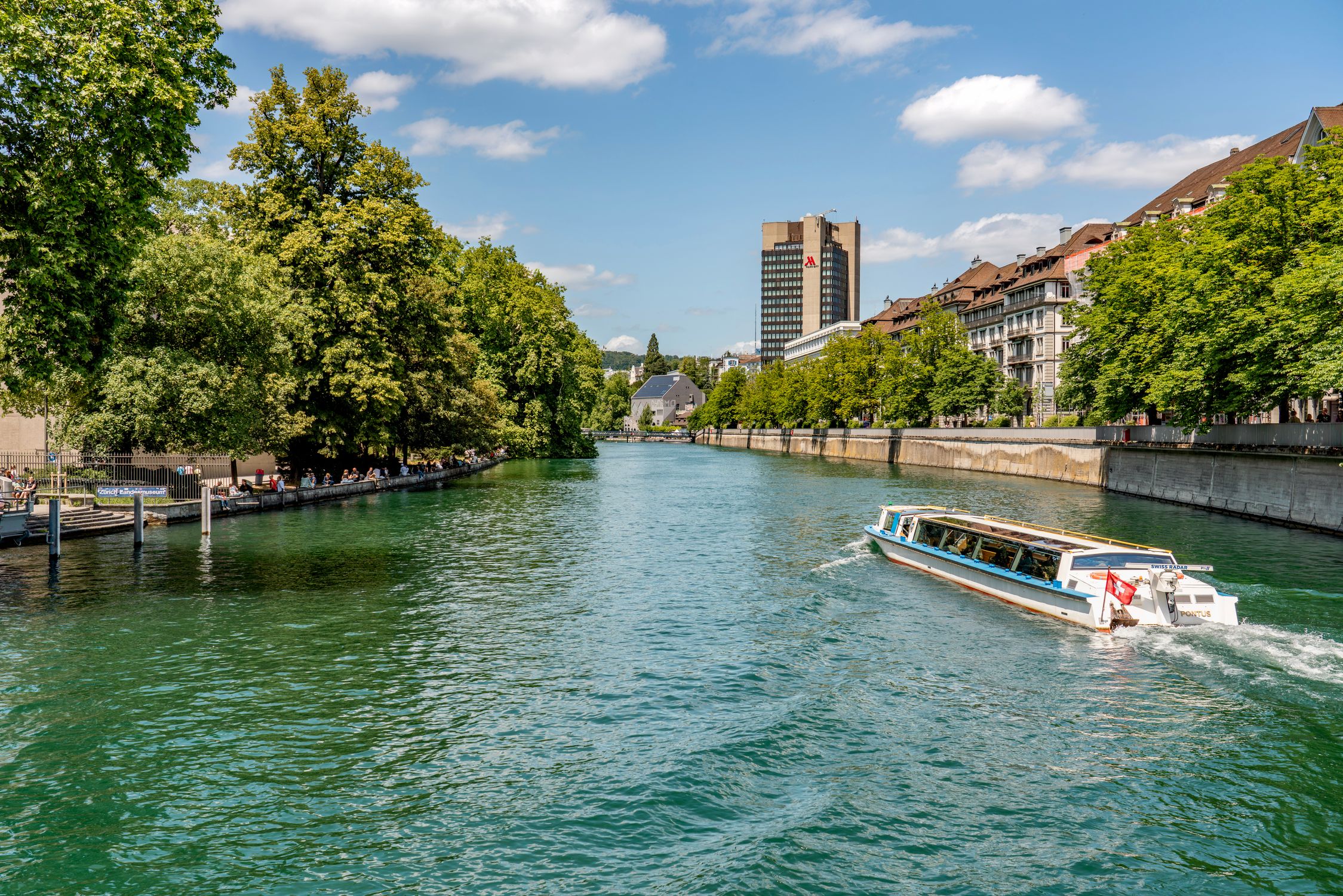 A tranquil view of a river in Zurich with a boat sailing under a blue, sunny sky.