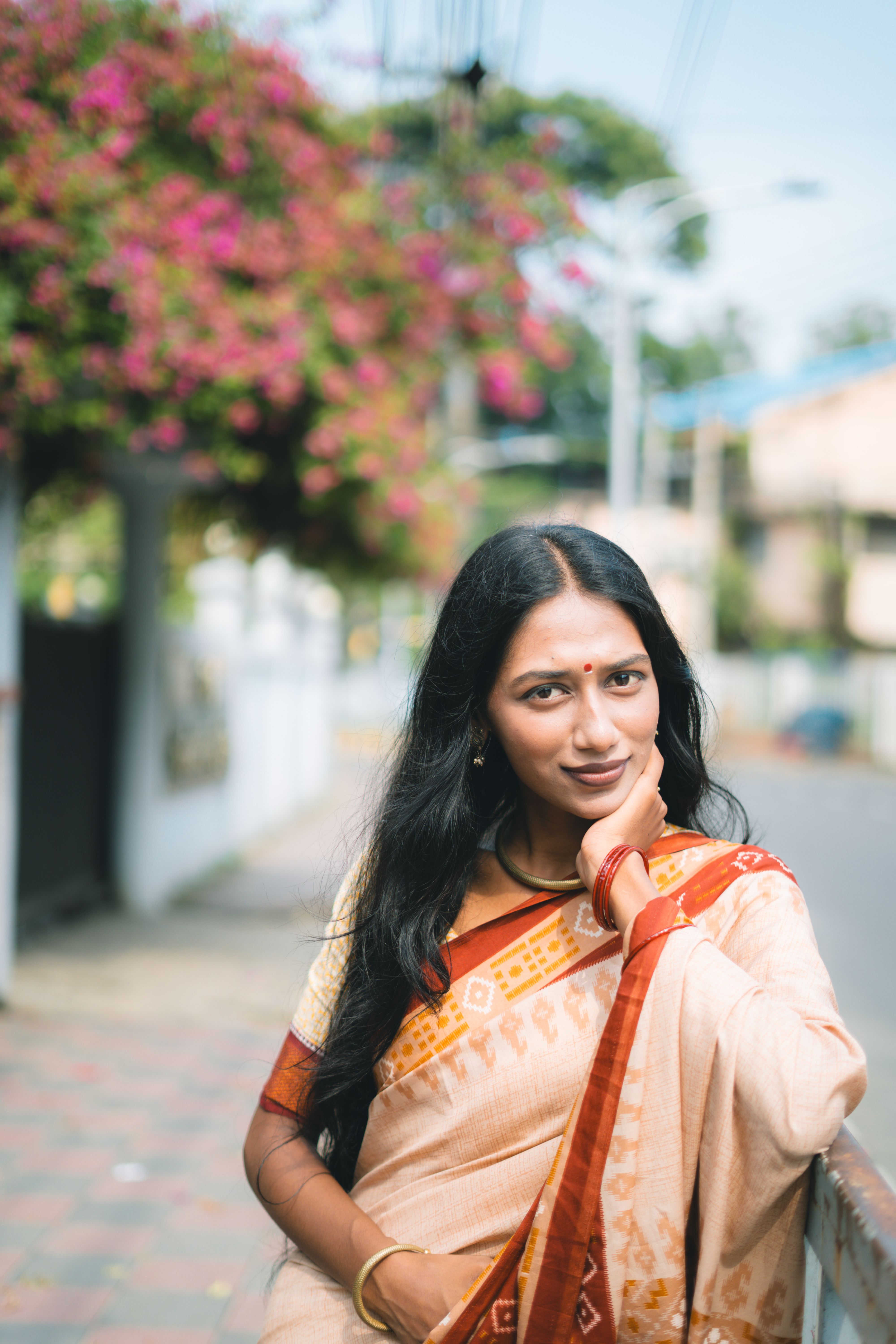 Woman poses in traditional attire outdoors, exuding confidence and elegance with a vibrant floral backdrop.
