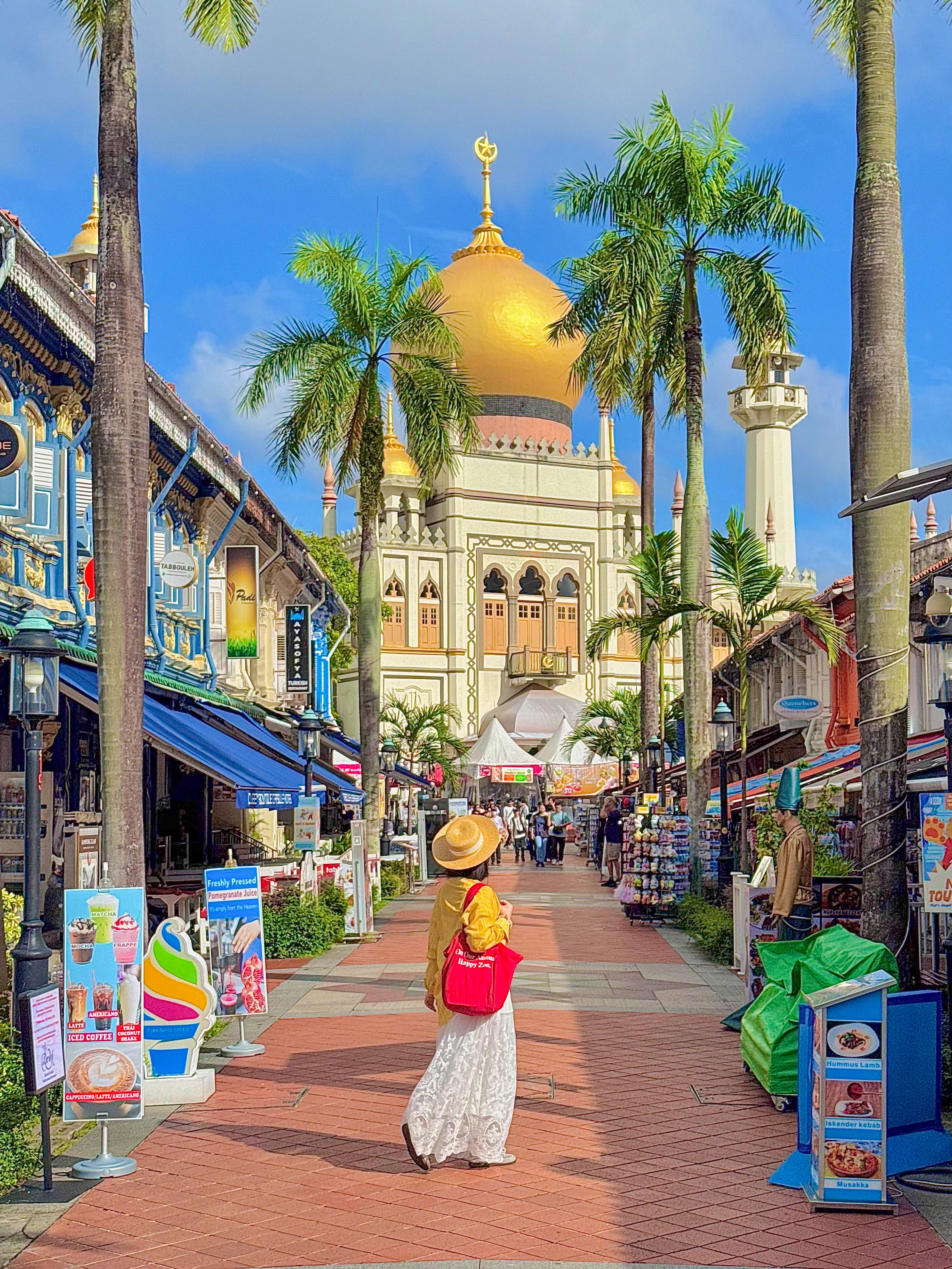 A woman in a sunhat and hijab strolls towards the iconic Sultan Mosque in Singapore, surrounded by vibrant shopfronts.