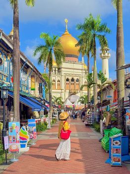 A woman in a sunhat and hijab strolls towards the iconic Sultan Mosque in Singapore, surrounded by vibrant shopfronts.